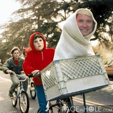 A man and a young boy riding bicycles on a road. The man is wearing a white hooded towel wrapped around his head and is smiling at the camera. The boy is also wearing a red hoodie and is holding onto the handlebars of the bicycle. The bicycle has a large metal basket attached to the front. In the background there are trees and a car parked on the side of the road. It appears to be a sunny day.