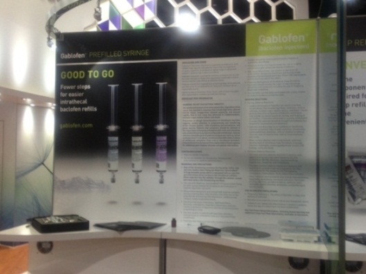 A trade show booth with a large banner on the left side. The banner has a black background with white text that reads "Gablofen Prefilled Syringe" and "Good to go". On the right side of the banner there is an image of three syringes with different sizes and colors. Below the syringes there are instructions on how to use the syringe. The booth has a white countertop and a black tray on the floor. In the background there appears to be a large screen with a colorful design.