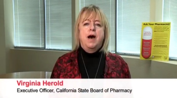 A screenshot from a video interview with Virginia Herold the Executive Officer of the California State Board of Pharmacy. She is standing in front of a window with blinds and there is a yellow sign on the right side of the image that reads "Ask Your Pharmacist". The woman is wearing a red blazer and a black top and she appears to be speaking into a microphone. She has blonde hair and is looking directly at the camera with a serious expression on her face.