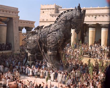 A large crowd of people gathered around a large statue of a horse. The statue is made of metal and appears to be made of wood or metal with intricate details and patterns. The horse is standing on its hind legs with its front legs stretched out and its head turned to the side. The crowd is gathered around the statue some of them are holding spears and shields while others are looking up at it. In the background there are several ancient buildings with columns and arches. The sky is blue and the overall atmosphere of the image is one of excitement and anticipation.