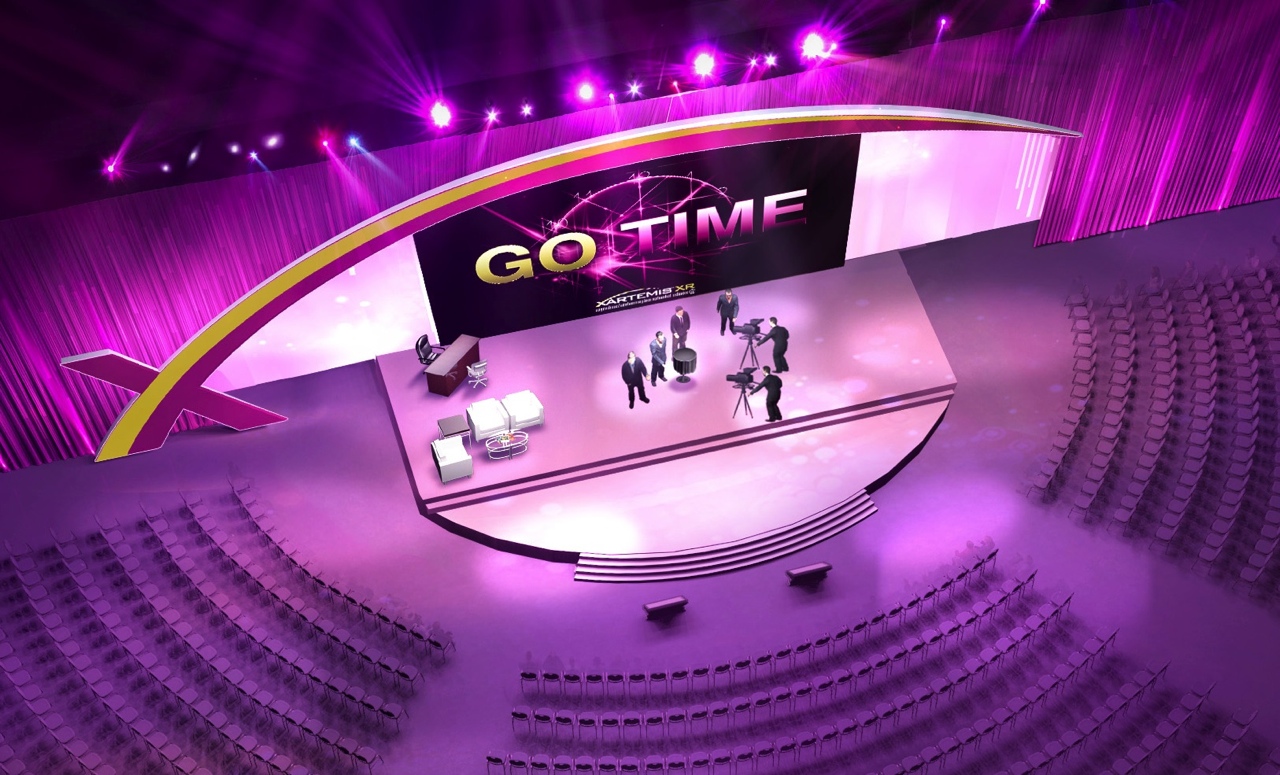 A 3D rendering of a stage set up for a concert or event. The stage is circular in shape and has a large screen in the center with the words "GO TIME" written in bold letters. The screen is surrounded by a pink and purple gradient background. On the left side of the stage there is a podium with a microphone and a few people standing on it. The audience is seated in rows of empty seats facing the stage. The overall color scheme of the image is purple and pink giving it a futuristic and energetic feel.