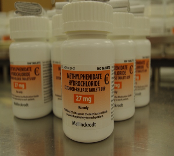 A row of white medicine bottles with orange labels. The bottles are arranged in a row on a metal countertop. The labels on the bottles read "Methylphenidate Hydrochloride Extended-Release Tablets USP 27 mg" and "Rx only". The bottles appear to be new and unused. The background is blurred but it seems to be a pharmacy or medical facility.