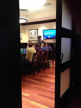 A group of people sitting on wooden chairs in a room with a wooden floor. The room appears to be a conference room or a meeting room as there is a large screen on the wall in the background. The people are gathered around a table with a laptop and other items on it and there are several framed pictures hanging on the walls. The image is taken from a high angle looking down on the people and the table. The overall mood of the image is casual and relaxed.