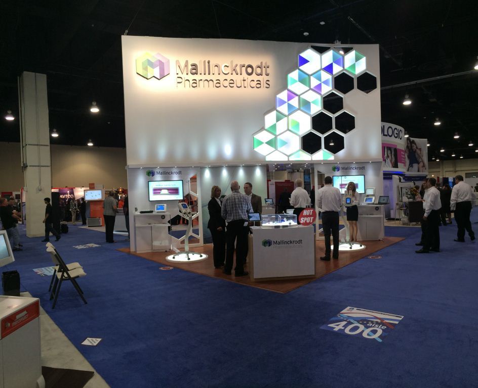 A trade show booth with a blue carpeted floor and white walls. The booth has a large white banner with the logo of Mallinckrodt Pharmaceuticals on the top left corner. The banner also has a colorful geometric design on the right side. In front of the booth there are several people standing and interacting with the booth. On the left side there is a desk with a computer monitor and other equipment. In the background there appears to be a large screen and other booths. The overall atmosphere of the image is busy and bustling.