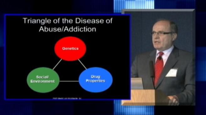 A slide from a presentation on the Triangle of the Disease of Abuse/Addiction. It shows a man standing at a podium and speaking into a microphone. He is wearing a suit and tie and appears to be giving a speech. The slide has a blue background with white text.<br /><br />The slide is divided into three sections. The first section is labeled "Genetics" the second section is titled "Social/Environment" and the third section is labeled "Drug Properties". Each section has a red circle with the word "Drug" written inside it representing the different types of drugs and their properties. The red circle represents genetics the green circle represents social/environment and the blue circle represents drug properties.