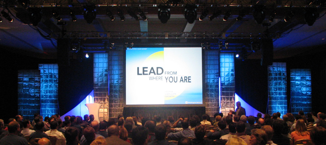 A large stage with a large screen in the center. The screen displays the words "LEAD FROM WHERE YOU ARE" in bold white letters. The stage is set up with blue lights and there is a podium on the right side of the stage. The audience is seated in rows of chairs facing the stage and attentively listening to the speaker. The background is dark and there are large windows on both sides of the screen allowing natural light to enter the room. The overall atmosphere of the image is professional and inviting.