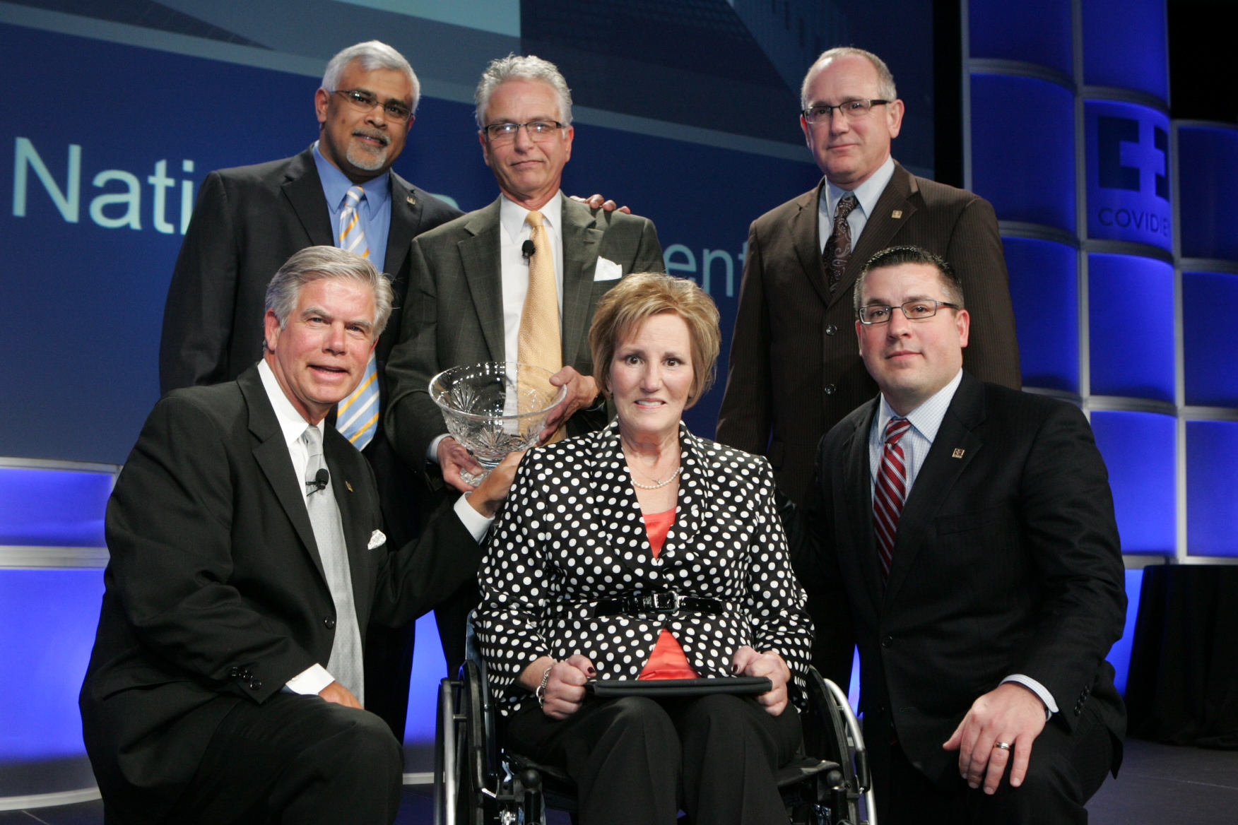 A group of six people posing for a photo on a stage. There are six men and one woman in the photo all dressed in formal attire. The woman is sitting in a wheelchair and is holding a large trophy. The man on the left is wearing a suit and tie and is smiling at the camera. The other six men are standing behind her also smiling. The background of the photo is blue with the words "National Geographic" and "COVIDIEN" written in white. The stage appears to be set up for a news conference or event.