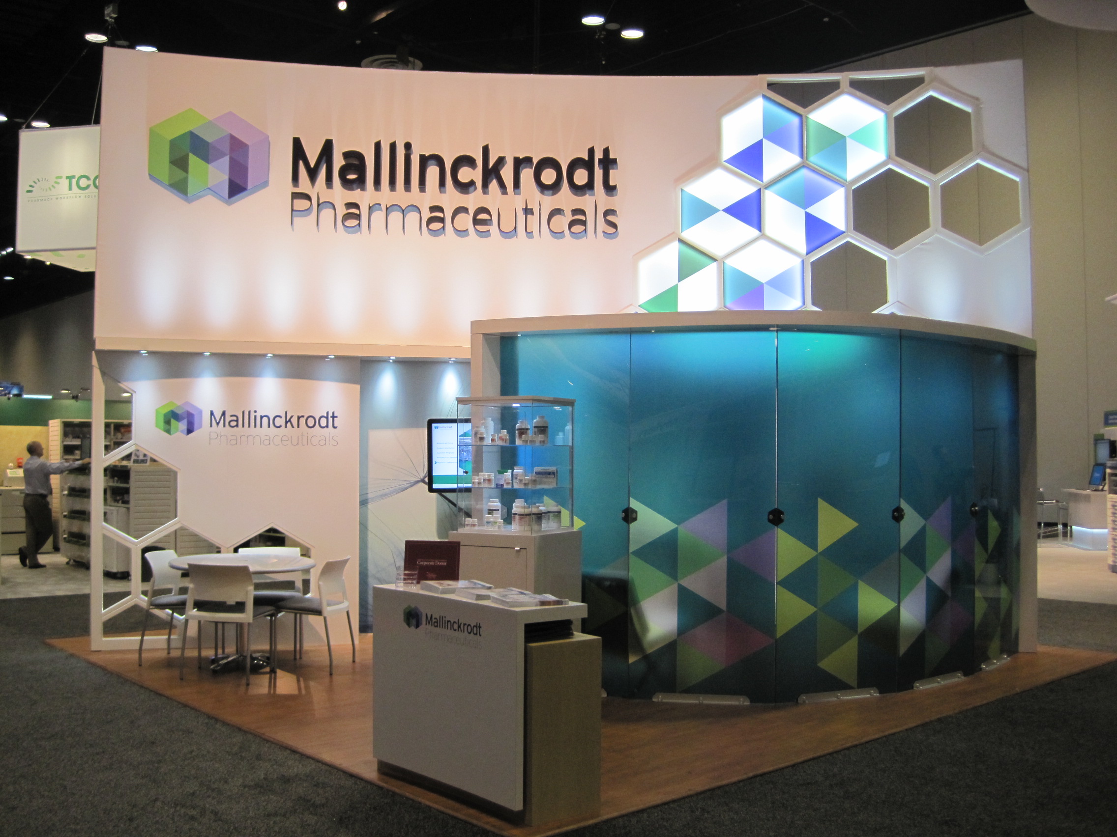 A trade show booth for Mallinckrodt Pharmaceuticals. The booth has a white background with the company's logo in blue and green hexagons. On the right side of the booth there is a large blue glass door with a geometric pattern. In front of the door there are two white tables and chairs with a computer monitor on top. There is also a small desk with a laptop and other items on it. The floor is covered with a gray carpet.