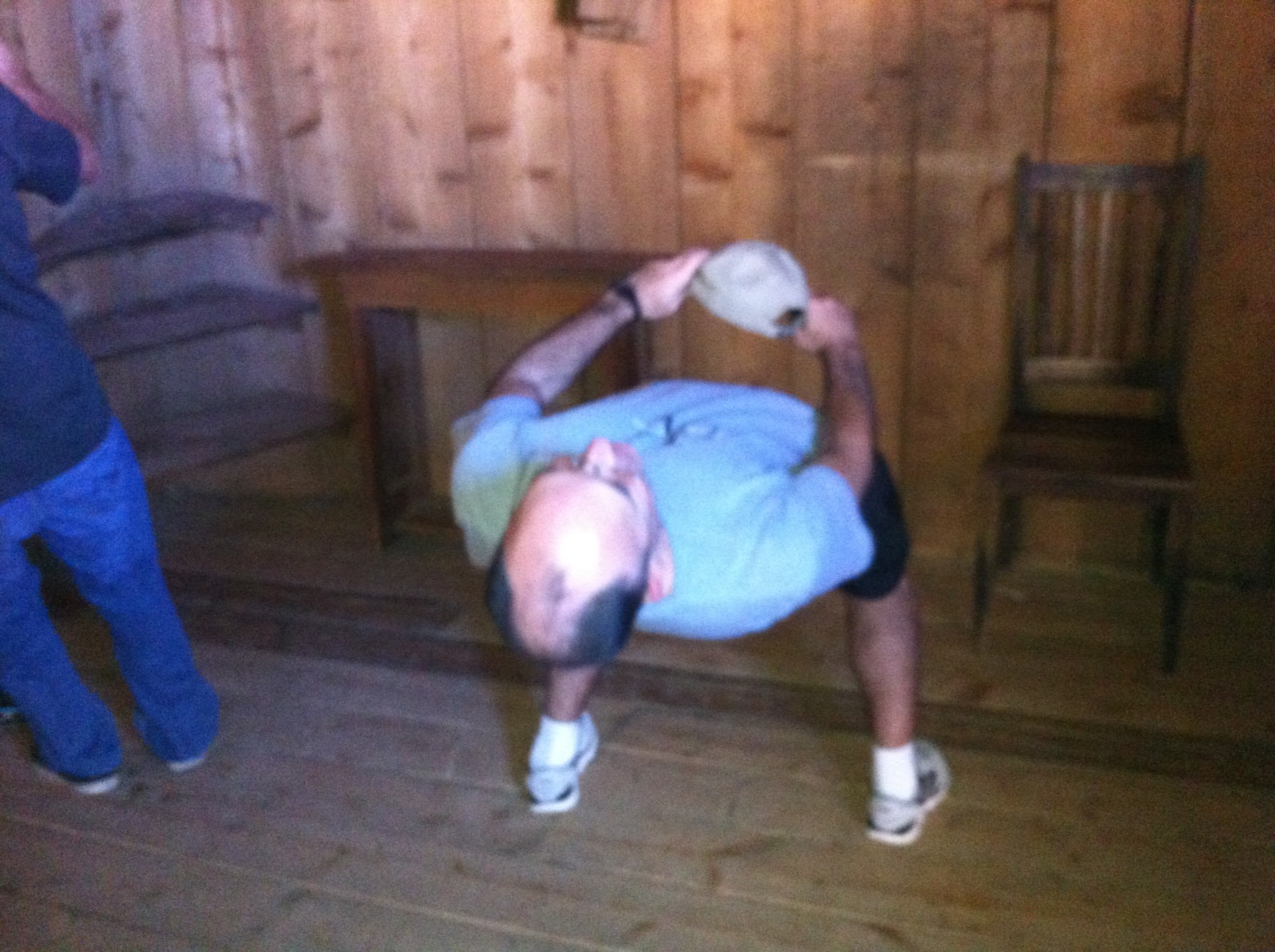 A man in a room with wooden walls and a wooden floor. He is in the middle of a handstand with his arms stretched above his head and his legs bent at the knees. He appears to be in the process of performing a push-up or handstand. The man is wearing a blue t-shirt black shorts and white sneakers. On the left side of the image there is another person standing and on the right side there are two wooden chairs. The image is taken from a low angle looking up at the man in the center of the frame.