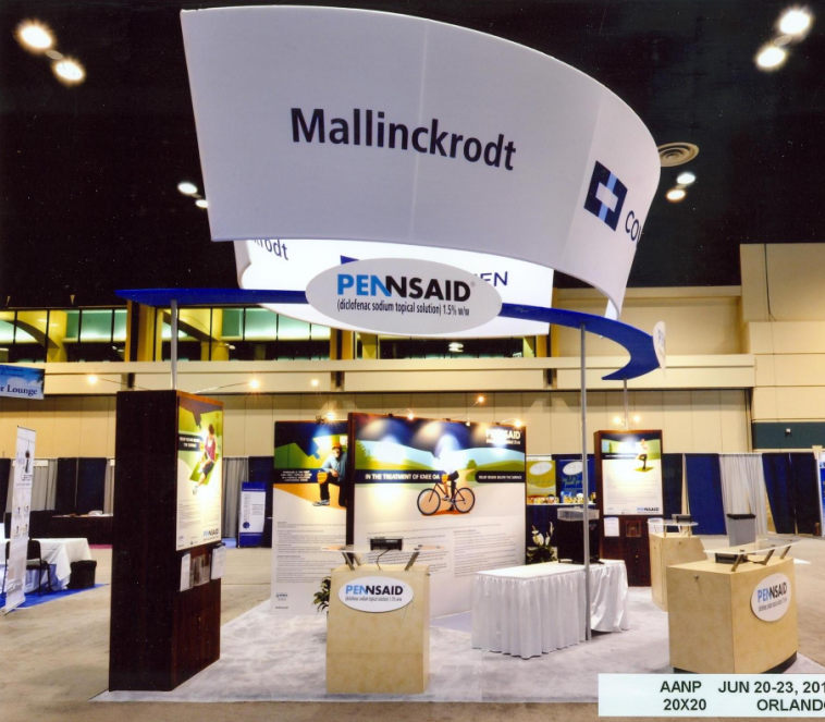 A trade show booth with a large white banner that reads "Mallinckrodt" and "Pennsaid" in blue letters. The booth is located in a large hall with a high ceiling and is decorated with blue and white banners. There are several tables and chairs set up in front of the booth and a few people can be seen in the background. On the left side of the image there is a large screen displaying information about the booth's services. The overall atmosphere of the trade show is modern and professional.