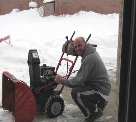 A man kneeling on the ground next to a snow blower. He is wearing a grey goodie black pants and black shoes. He has a big smile on his face and is holding the handle of the blower with both hands. The blower is red and black in color and has a large snow plow attached to it. The man is kneeling in front of a brick building with snow on the sidewalk and a fence in the background. The sky is overcast and the ground is covered in snow.