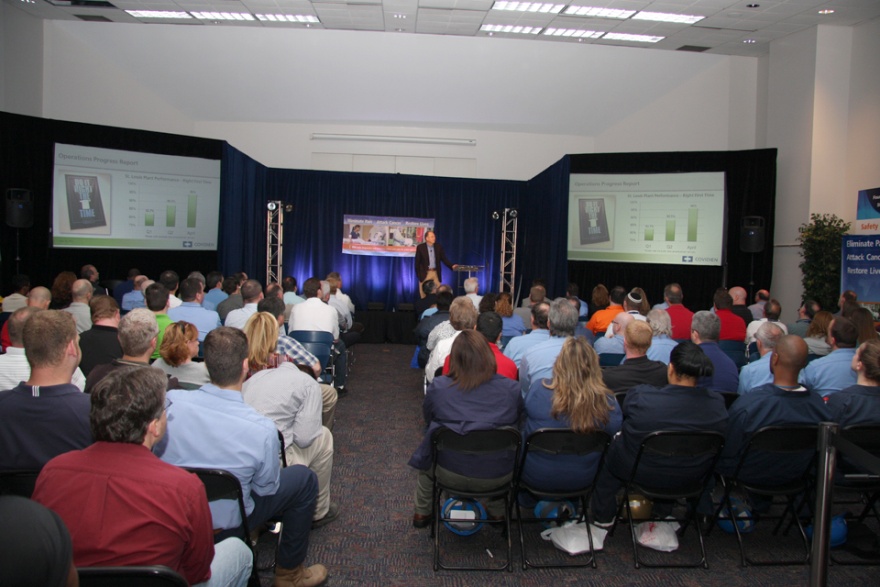 A large conference room with a stage in the center. The stage is set up with a blue curtain and there are two large screens on either side of the stage. On the stage there is a man standing at a podium and speaking into a microphone. He appears to be giving a presentation. The audience is seated in rows of chairs facing the stage and attentively listening to the speaker. The room has a high ceiling with recessed lighting and a carpeted floor. There is a banner on the right side of each room with text on it.