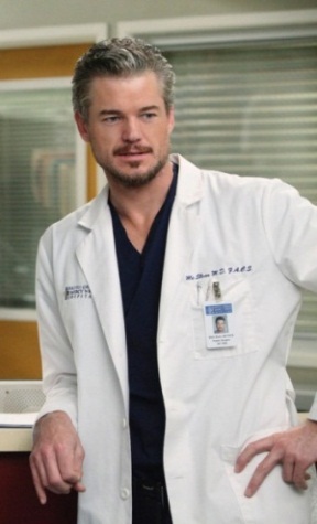 A man standing in a hospital room. He is wearing a white lab coat with a name tag that reads "Dr. David Siegel" and a blue scrub top underneath. He has a serious expression on his face and is looking directly at the camera. The background shows a window with blinds and a desk with a computer monitor. The man appears to be in his late twenties or early thirties.