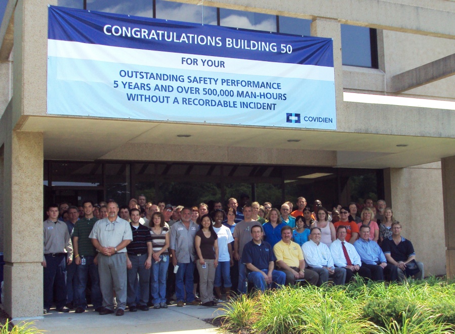 A group of people standing in front of a building with a large blue banner that reads "Congratulations Building 50 for your outstanding safety performance 5 years and over 500000 man-hours without a recordable incident". The group is composed of men and women of different ages and ethnicities and they are all smiling and posing for the photo. The building appears to be a modern structure with a concrete facade and large windows. The sky is blue and there are trees and bushes in the background.