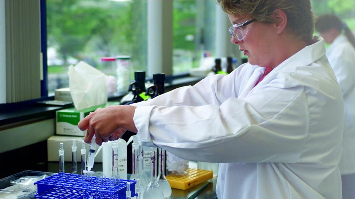 A young woman in a laboratory setting. She is wearing a white lab coat and glasses and is standing in front of a workbench with various laboratory equipment. On the workbench there are several test tubes flasks and other scientific equipment. The woman is holding a pipette and appears to be conducting a scientific experiment. In the background there is another person working in the same lab. The laboratory has large windows allowing natural light to enter the space.