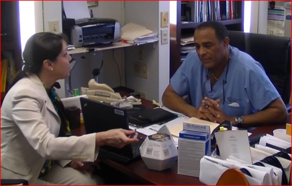 A man and a woman sitting at a desk in an office. The man is wearing a blue scrub suit and a stethoscope around his neck. He is sitting in front of a computer monitor and appears to be engaged in a conversation with the woman. The woman is sitting next to him and is gesturing with her hand as if she is explaining something to him. On the desk there are various office supplies such as a laptop a telephone and a stack of papers. There is also a printer and a bookshelf in the background. The image appears to have been taken during the day.