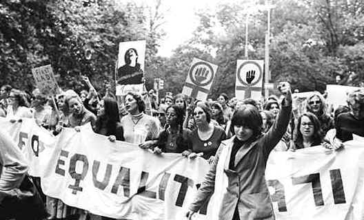 A black and white photograph of a group of people participating in a protest or rally. The people are holding banners and placards with various messages written on them. The banner reads "EQUALITY" in bold letters and there is a woman in the center of the group with her arms raised in the air. She is wearing a suit and appears to be leading the group. The background shows trees and a building suggesting that the protest is taking place outdoors. The image appears to have been taken in a park or a wooded area.