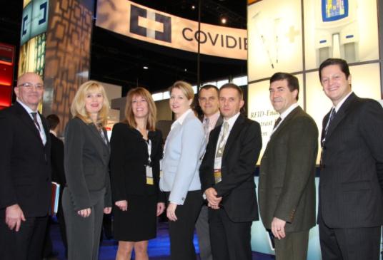 A group of seven people standing in front of a booth at a trade show. The booth has a blue carpet and a large banner that reads "COVIDIEN" in white letters. The people in the group are dressed in formal business attire and are smiling at the camera. On the left side of the image there is a man wearing a suit and glasses and on the right side there are two men wearing suits and ties. In the center of the group there appears to be a woman wearing a black dress and a man in a suit. Behind them there seems to be another booth with a sign that says "Covidien". The background is dark suggesting that the photo was taken at night.