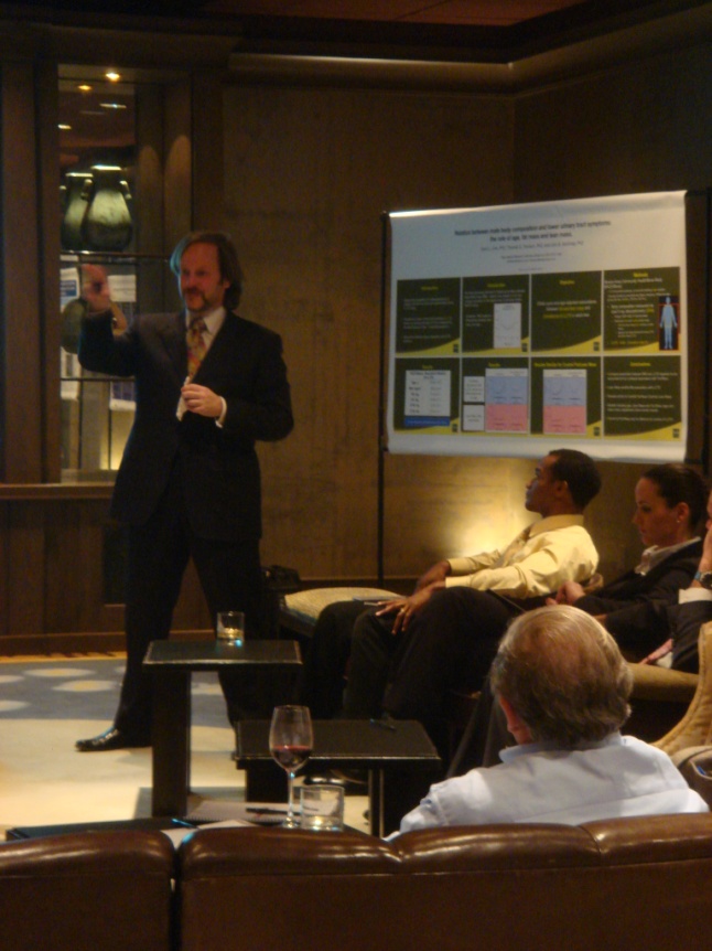 A man standing in front of a group of people in a conference room. He is wearing a suit and tie and appears to be giving a presentation. The room has a wooden paneled wall and a large screen on the right side of the image. The people in the room are sitting on leather sofas and are attentively listening to the man speak. There is a small table in the center of the room with a glass of wine on it.