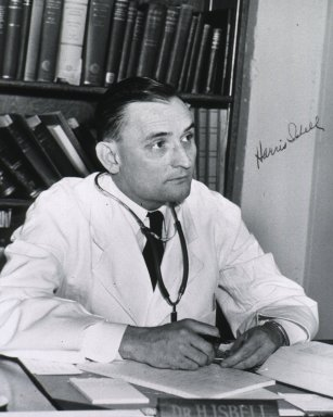 A black and white photograph of a man sitting at a desk in an office. He is wearing a white lab coat and a stethoscope around his neck. He appears to be a doctor or a medical professional as he is holding a pen and writing on a piece of paper. The desk is cluttered with papers books and other medical supplies. Behind him there is a bookshelf filled with books. The man is looking off to the side with a serious expression on his face. The photograph is signed by the man in the photograph "Dr. Hirschfeld."