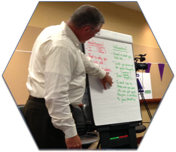 A man standing in front of a whiteboard in a conference room. He is wearing a white shirt and appears to be writing on the whiteboard with a marker. The whiteboard is mounted on a black easel and there is a camera on a tripod next to it. The man is pointing to something on the board with his right hand and is looking down at it with a focused expression on his face. The background of the image is a beige wall with a purple banner hanging on it.