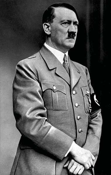 A black and white portrait of a man in a military uniform. The man is standing with his hands in his pockets and is facing the camera. He is wearing a blazer with a high collar and a tie. The blazer has several buttons and a badge on the left side of his chest. He has a mustache and is looking off to the side with a serious expression on his face. The background is plain and dark making the man the focal point of the image.