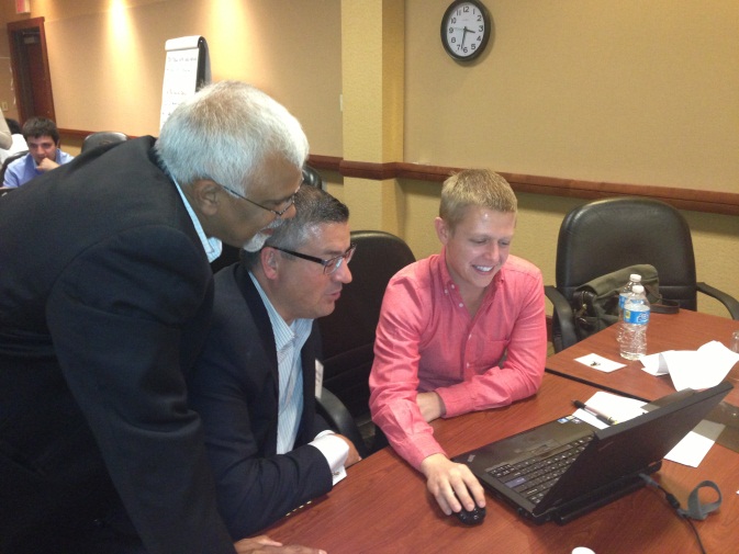 Three men sitting at a conference table with a laptop in front of them. The man on the left is an older man with white hair and glasses wearing a black suit. He is leaning over the table and appears to be explaining something to the young man in the middle who is wearing a pink shirt. The young man is smiling and looking at the laptop screen. There are water bottles and papers on the table and a clock on the wall in the background.