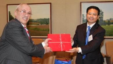 Two men in a room with a painting on the wall in the background. The man on the left is an older man with white hair and glasses wearing a suit and tie. He is holding a red gift box with a white ribbon tied around it. The other man is a younger man with dark hair and a blue tie. Both men are smiling and appear to be presenting the gift to the older man. There is a blue bag on the floor next to them.