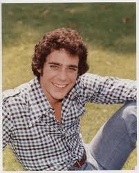 A portrait of a young man sitting on a grassy field. He is wearing a black and white checkered shirt and blue jeans. He has curly brown hair and is smiling at the camera. His arms are stretched out to the sides and his legs are slightly bent at the knees. The background is blurred but it appears to be a sunny day with trees and greenery. The overall mood of the image is relaxed and casual.