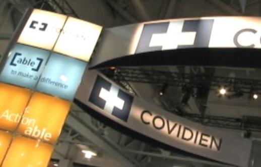 A large signboard with the logo of Covidien a pharmaceutical company on it. The signboard is located in an indoor space with a high ceiling and metal beams. The logo is a white cross with the word "COVIDIEN" written in black letters. Below the logo there is a smaller signboard that reads "Cable" and "Actionable". On the left side of the signboard there are several smaller signs that read "Table 7 to make a difference". The overall color scheme of the image is blue orange and white.