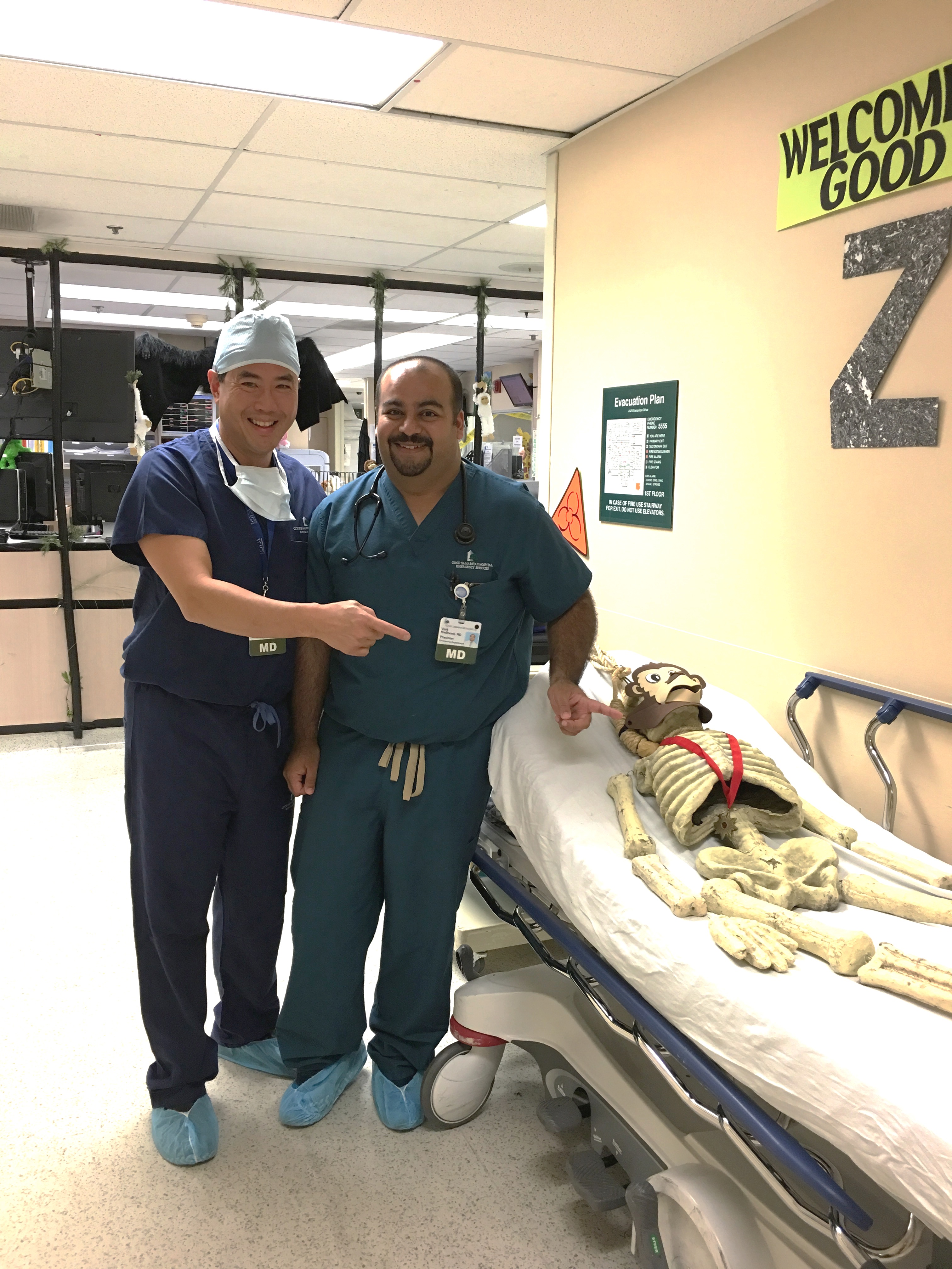 Two men in scrubs standing next to a hospital bed with a skeleton on it. The man on the left is wearing a blue scrub suit and a white cap and he is pointing at the camera with a smile on his face. He is also wearing a stethoscope around his neck and has a name tag attached to his shirt. On the right side of the image there is a sign that reads "Welcome Good Z" and on the wall behind the bed there are medical equipment and a sign with the letter "Z" on it indicating that this is a hospital room.