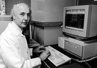 A black and white photograph of a man sitting at a desk in front of a computer. The man is wearing a lab coat and appears to be in his late 60s or early 70s. He has a serious expression on his face and is looking directly at the camera.<br /><br />On the desk there is a computer monitor a keyboard and a CPU. The computer monitor is turned on and the keyboard is placed next to it. The CPU is also turned on. The desk is cluttered with wires and other medical equipment. The background of the image is blurred but it seems to be a laboratory or laboratory setting.