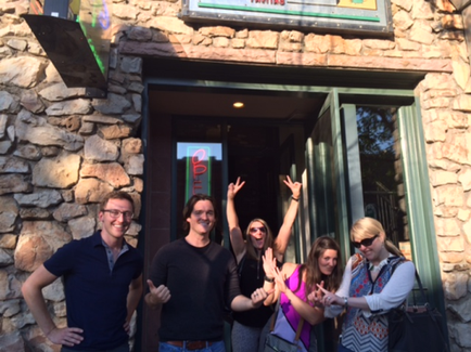 A group of five young people standing in front of a stone building with a sign that reads "came". They are all smiling and making a peace sign with their hands. The building appears to be a restaurant or came as there is a sign above the entrance with the restaurant's name on it. The people are standing outside the building and there are trees visible in the background.
