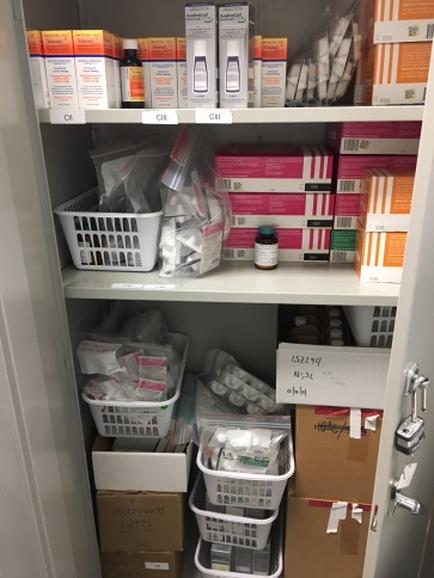 A white cabinet with three shelves. On the top shelf there are several boxes of different sizes and colors including orange pink and white. The boxes are arranged neatly on the shelves with some of them open and some closed. There are also two white plastic baskets on the bottom shelf which are filled with medical supplies such as bandages bandages in plastic bags and a small bottle of medicine. The bottom shelf has several cardboard boxes with labels on them and there is a lock on the right side of the cabinet. The cabinet appears to be in a room with a white wall and a door.
