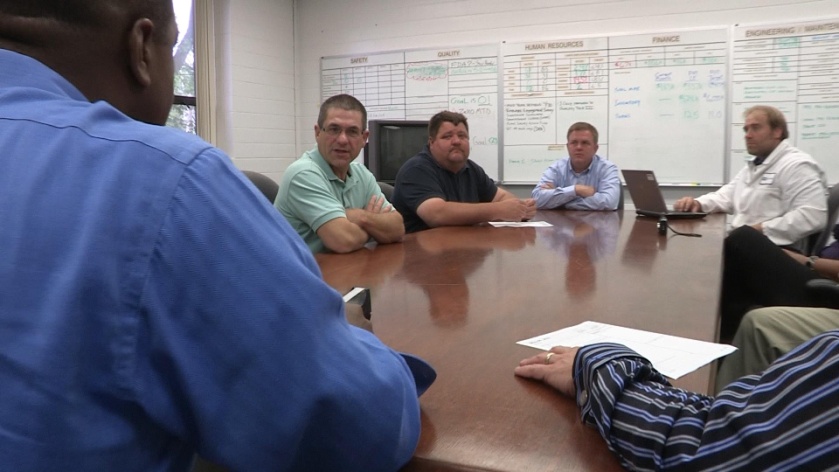 A group of six men sitting around a wooden conference table in a meeting room. They are all facing the same direction and appear to be engaged in a discussion. The man on the left is wearing a blue shirt and is standing in front of the table while the others are sitting in chairs around the table. On the right side of the image there is a man wearing a white lab coat and a woman wearing a striped shirt. In the background there are whiteboards and a projector screen. The room appears to be well-lit with natural light coming in from the windows.