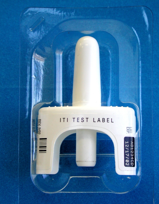 A clear plastic packaging with a white test tube inside. The test tube has a label on it that reads "ITI TEST LABEL" and has a barcode on the bottom right corner. The packaging appears to be sealed and is placed on a blue background.