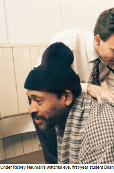 A man wearing a black beanie and a striped shirt. He has a beard and is looking to the side with a slight smile on his face. Behind him there is another man who is leaning over and appears to be helping him. The background is a white tiled wall. The image appears to have been taken in a room with a window.