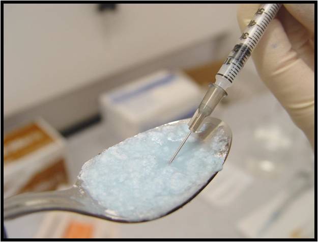 A close-up of a person's hand holding a syringe with a needle inserted into a spoon. The syringe is filled with a light blue substance which appears to be sugar or a similar substance. The spoon is silver and has a curved handle. The background is blurred but it seems like the person is in a laboratory setting with other medical equipment visible.