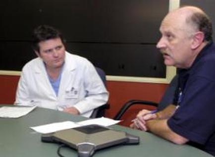 Two men sitting at a table in a meeting room. The man on the left is wearing a white lab coat and appears to be a doctor or a medical professional. He is looking at the other man with a serious expression on his face. On the table in front of them there are papers and a calculator. The background of the image is a blackboard and a red wall. It appears that the two men are engaged in a conversation.