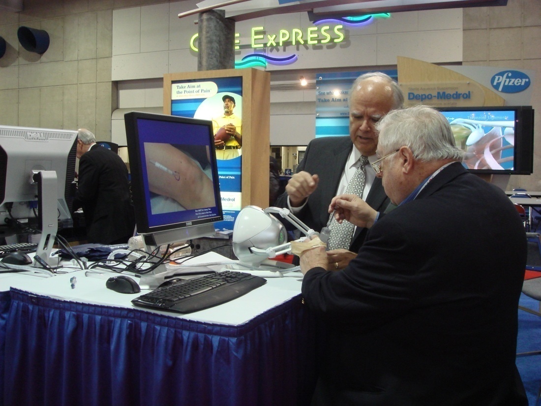 Two men standing in front of a desk with multiple computer monitors and keyboards. The desk is covered with a blue tablecloth and there is a sign that reads "Express" above it. The man on the left is wearing a suit and tie and appears to be explaining something to the man in the middle who is holding a white object in his hand. On the right side of the desk there is another man wearing a black suit and glasses. In the background there are several other people and a large screen displaying an image of a person. The image appears to have been taken at an exhibition or trade show.