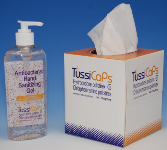 A bottle of TussiCaps Antibacterial Hand Sanitizing Gel and a box of tissues. The bottle is transparent and has a pump dispenser on top. The box is white with orange and blue accents. The label on the bottle is orange and white with the brand name "TussiCaps" written in bold black letters. The tissues inside the box are white and appear to be made of tissue paper. The background is a solid blue color.