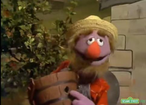 A still from the Sesame Street show. It shows the character Elmo holding a large clay pot. Elmo is wearing a yellow straw hat and a red scarf around his neck. He has a big smile on his face and is looking directly at the camera. The background shows a brick wall and a tree. The overall mood of the image is cheerful and playful.