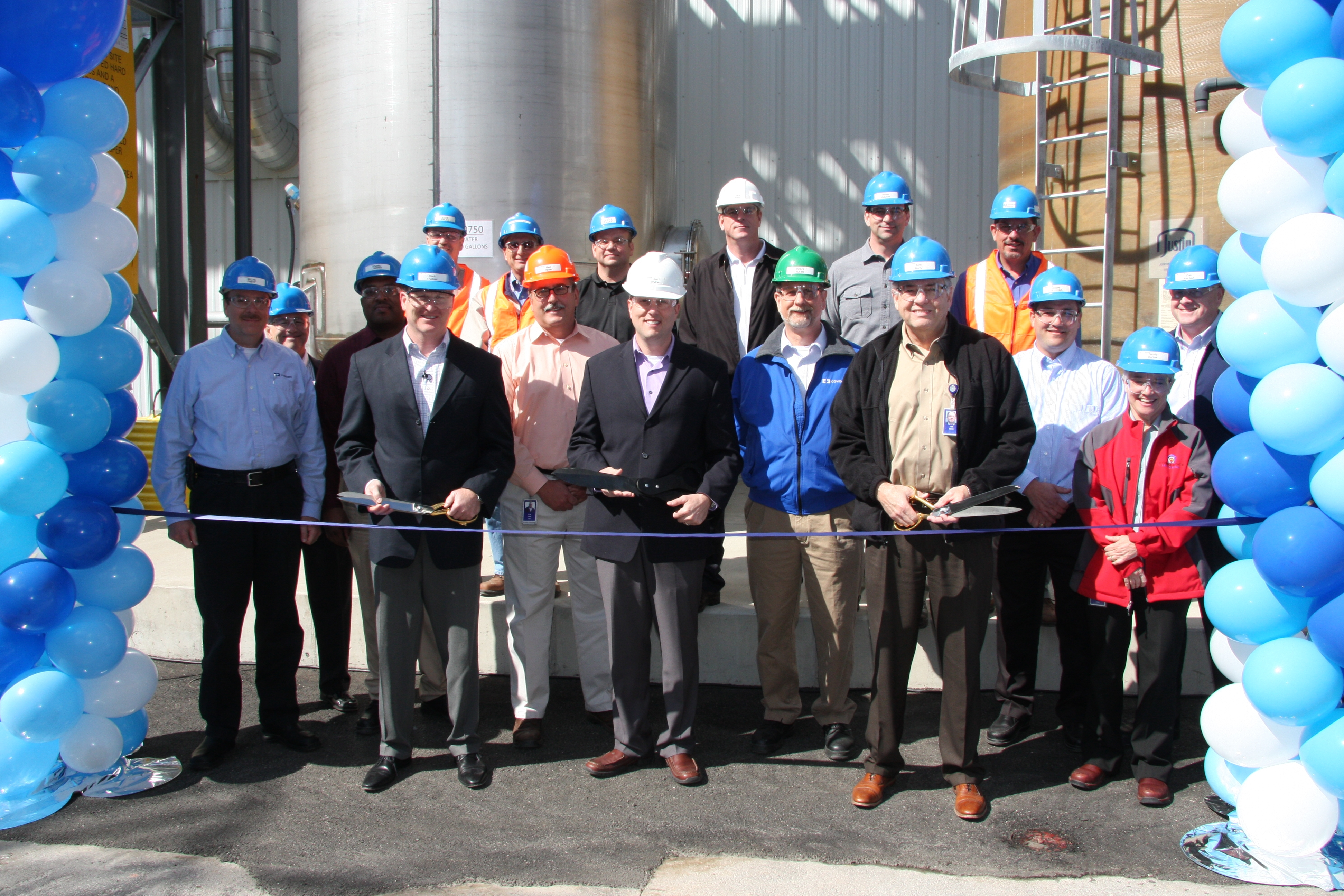 A group of people standing in front of a large industrial building with blue and white balloons on either side. They are all wearing hard hats and are holding a pair of scissors ready to cut the ribbon that is being cut. The group appears to be at a ribbon cutting ceremony as there is a large metal silo in the background. The sky is blue and the ground is wet suggesting that it has recently rained. The people in the group are smiling and looking at the camera.