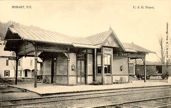 A black and white photograph of a train station in Hobart New York. The station is a two-story building with a sloping roof and a covered porch. The building is made of wood and has a sign that reads "U.S. & D. Depot." There is a bicycle parked on the platform in front of the building. The sky is overcast and there are trees in the background. The photograph appears to be from the early 20th century.