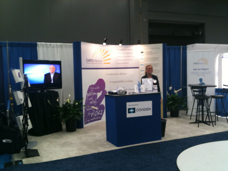 A trade show booth for the CARESAlliance sponsored by Covidien. A young woman is standing behind a blue table while a video presentation is playing on a flat screen television (to her right). The space includes signage tables chairs literature and potted plants. The primary color scheme is blue and white.