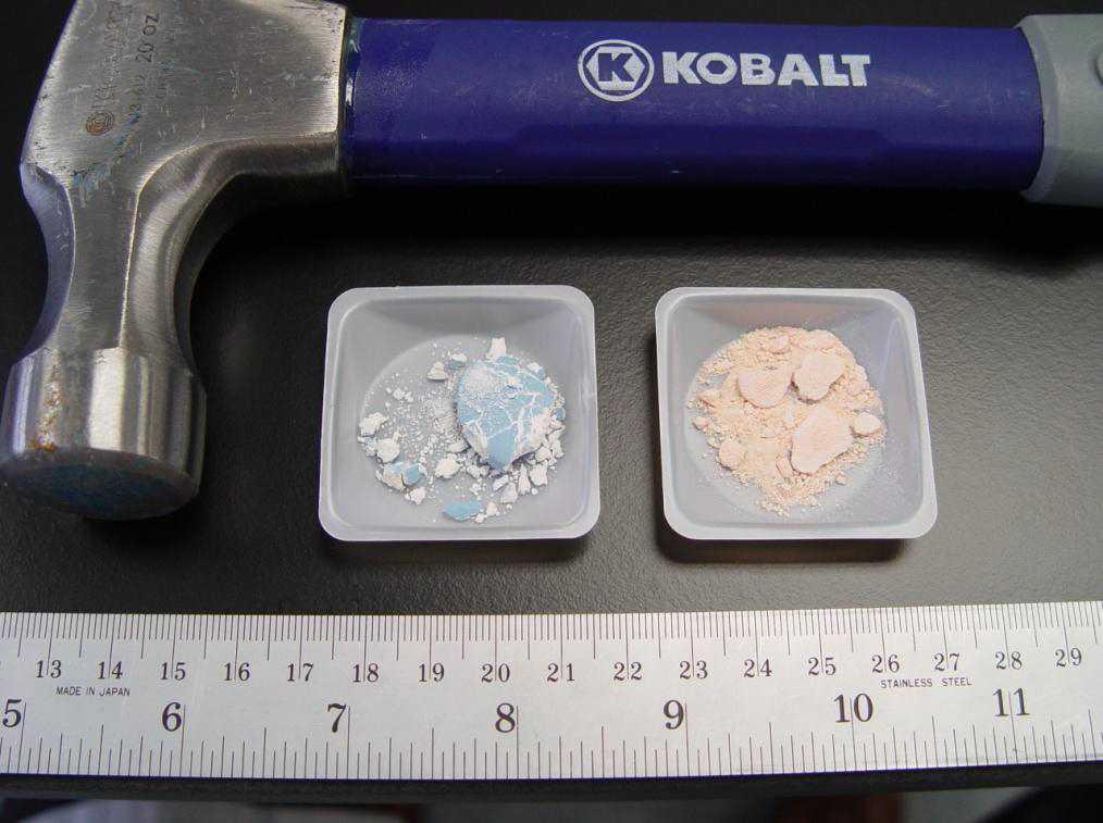 A Kobalt hammer with a blue handle and a silver head. The hammer is resting on a black surface with two small plastic containers next to it. The container on the left contains a small amount of white powder while the container in the middle contains a larger amount of pink powder. A ruler is also visible next to the hammer showing the scale of the scale. The background is blurred but it appears to be a desk or table.