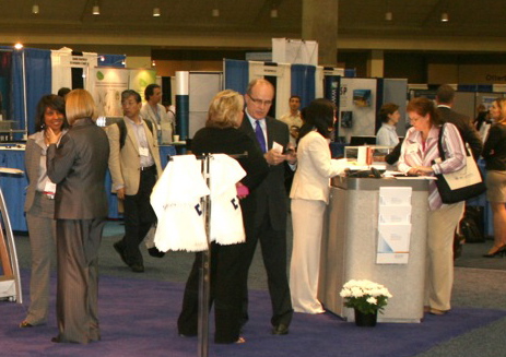 A group of people at an exhibition or trade show. There are several people in the image including a man in a suit and a woman in a white dress who appear to be engaged in conversation. They are standing in front of a booth with a sign that reads "Welcome" and there are several other booths in the background. The booth is decorated with blue and white banners and there is a vase of white flowers on the floor. The people are dressed in formal business attire and some are carrying bags. It appears to be a busy and bustling event.