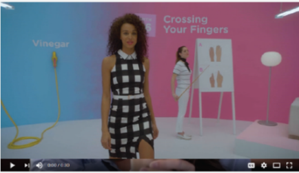 A screenshot from a video titled "Crossing Your Fingers". It shows a young woman with curly hair wearing a black and white checkered dress. She is standing in front of a pink background with the word "Vinegar" written in white. On the right side of the image there is a white easel with a diagram of the fingers on it. The woman is holding a yellow skipping rope and appears to be in the middle of a dance move.