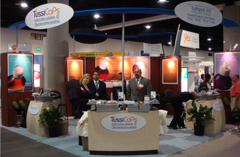 A trade show booth with a blue and orange color scheme. The booth has a sign that reads "TussiCaps" in white letters. There are several people standing around the booth some of them are looking at the display. In the background there are several other booths with orange and white signs and banners. There is also a man in a suit and tie standing in front of one of the booths. The overall atmosphere of the booth is professional and inviting.