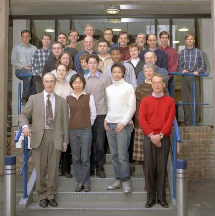 A group of people standing on the steps of a building. There are around 20 people in the group all of them are smiling and looking at the camera. They are arranged in a semi-circle with some standing on top of the stairs and others on the ground floor. The building appears to be a modern structure with large windows and a blue railing on the right side. The people are dressed in casual clothing and appear to be of different ages and ethnicity's. The image appears to have been taken in the early 20th century.